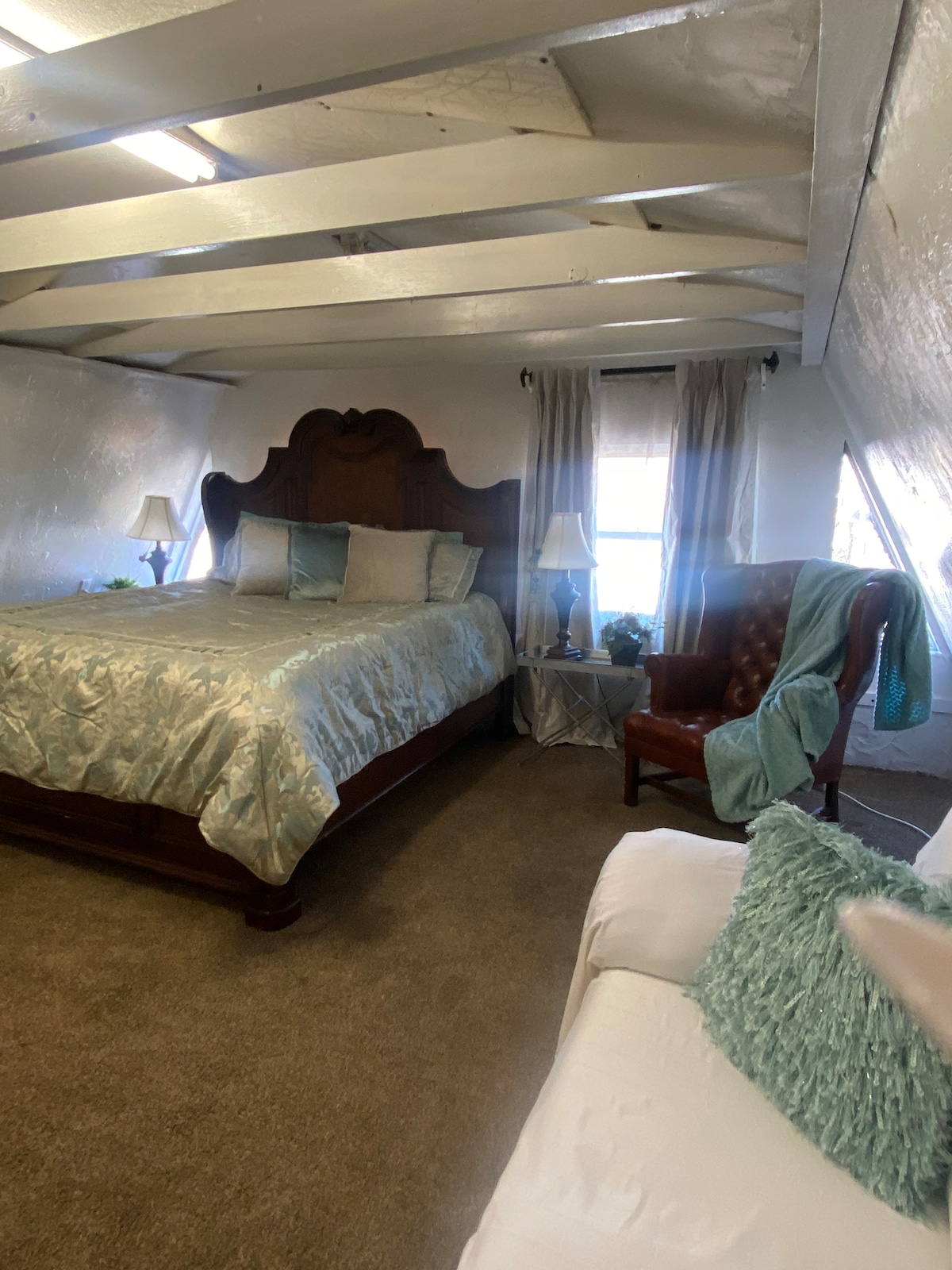 A spacious bedroom features a large bed with a light blue quilt, flanked by two bedside lamps. A vintage armchair sits in one corner, and a white couch is displayed opposite the bed. Natural light enters through window treatments that complement the soft color palette.