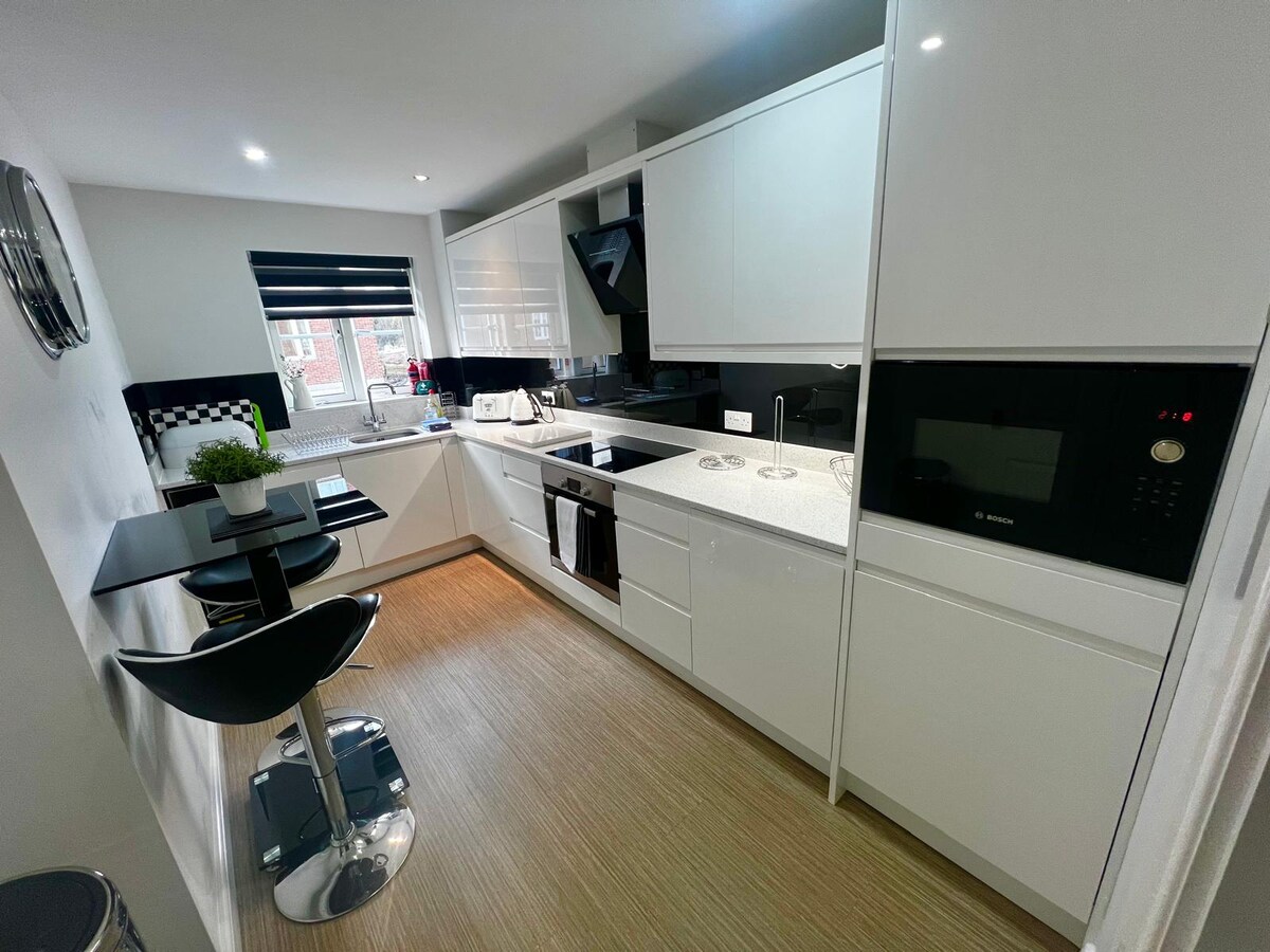 A sleek, modern kitchen features white cabinetry and stylish countertops. Equipped with a built-in electric oven, microwave, and fridge, the space includes a small breakfast bar with two high-backed stools. Natural light filters in from a window adorned with black blinds.
