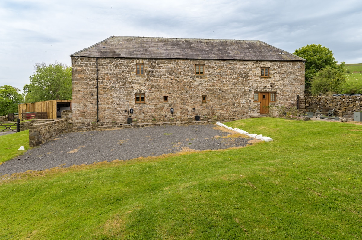 The exterior of a historic stone barn is showcased, featuring weathered stone walls and a wooden door. A spacious gravel area is visible in front, surrounded by green lawn. A small gravel path leads up to the entrance, with trees and countryside in the background.