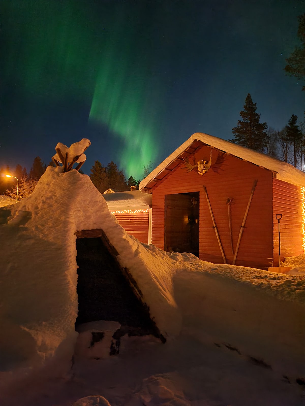 A rustic building, adorned with strings of warm lights, stands under a vibrant display of the Northern Lights. Snow-covered structures and trees frame the scene, while a snow mound with a wooden sculpture is positioned in the foreground, adding unique character to the landscape.
