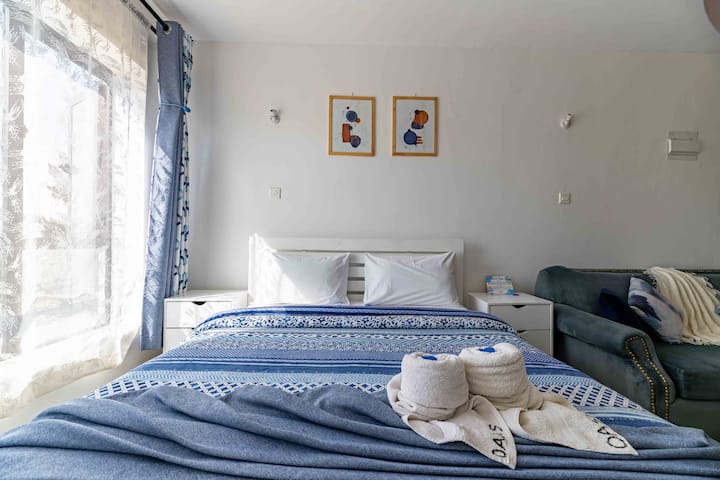 להתרווח בנוחות על מיטה מרווחת עם מצעים לבנים נקיים ושמיכות חמימות, מוקפת בגווני כחול מרגיעים ואור שמש חם שזורם דרך הווילונות השקופים.