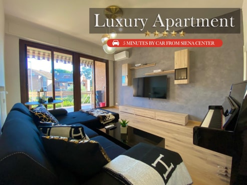 A modern living area features a spacious black sofa, paired with decorative cushions. A large television is mounted on a wall unit, and natural light filters in through sliding glass doors leading to an outdoor terrace. A piano is positioned in the corner.