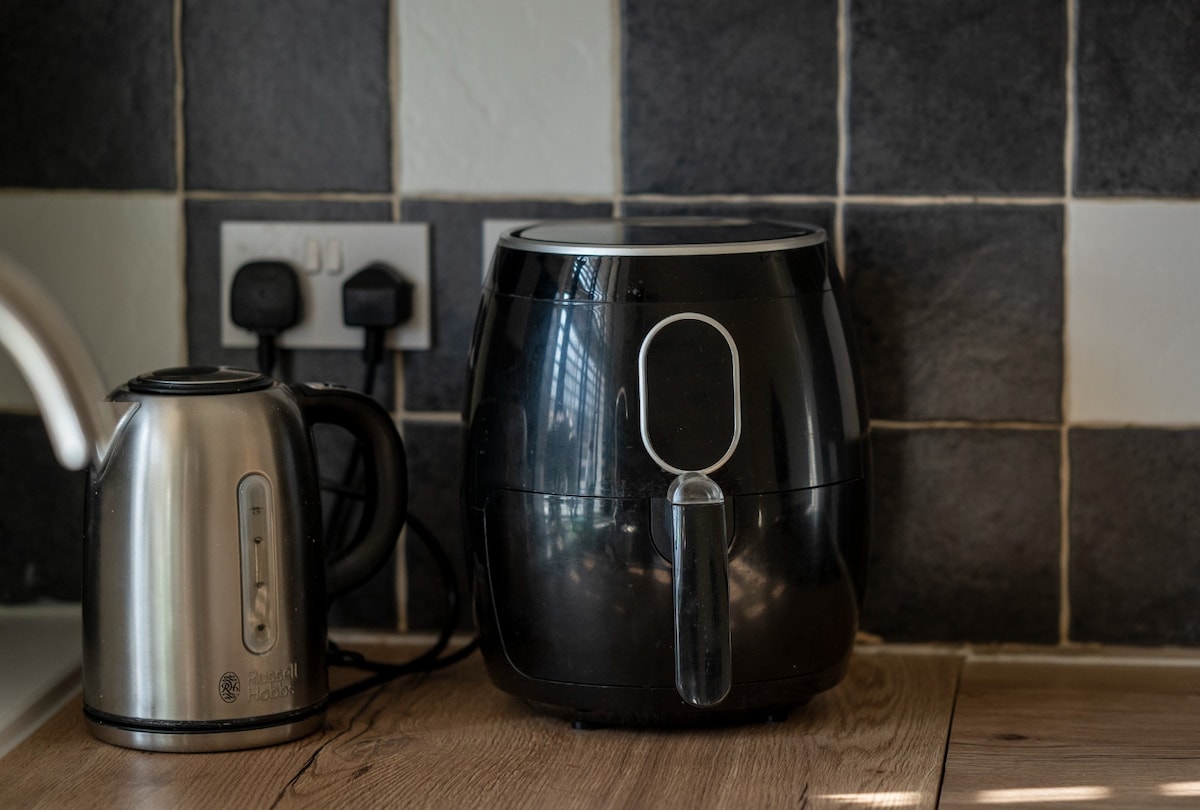 A black air fryer sits on a wooden countertop next to a stainless steel kettle. The background features a tiled wall with alternating black and white tiles, enhancing the kitchen's modern design. Electrical outlets are visible, accommodating the appliances.