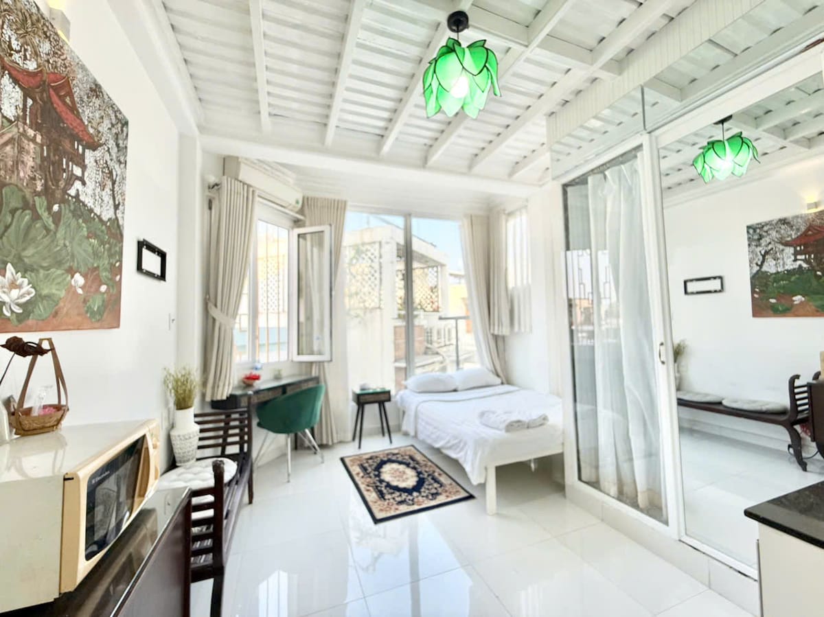 A bright sleeping area features a large window allowing natural light to fill the space. Light-colored walls are complemented by decorative wall art, and a spacious bed is positioned beside a wooden chair and table. A mirror reflects the room's open layout.