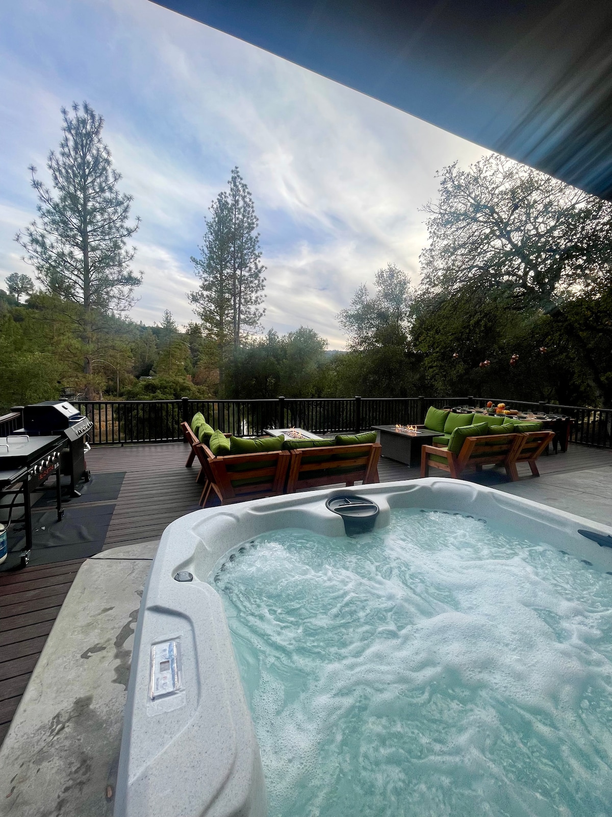 A hot tub is positioned on a spacious deck with jets bubbling, surrounded by outdoor lounge furniture in vibrant green. A propane grill stands nearby, framed by tall trees and a sky with gentle clouds, enhancing a relaxed atmosphere for outdoor enjoyment.
