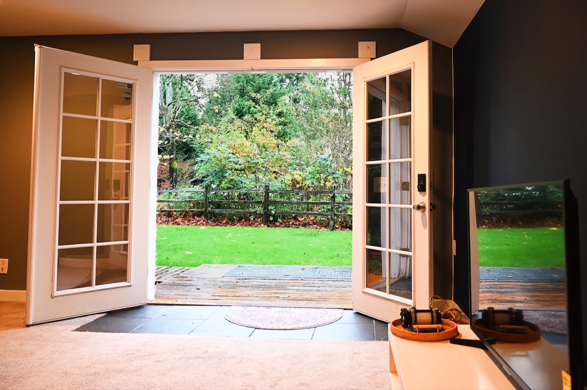 Double French doors open to a lush green yard, framed by trees, providing a seamless connection to nature. A smooth indoor surface leads to the outdoor deck, while a cozy living room area includes a television and decorative elements.