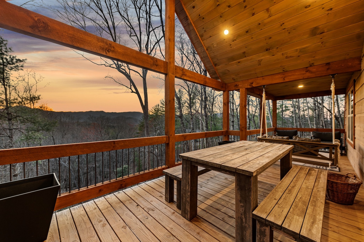 A spacious deck features wooden furniture, including a large table and benches, surrounded by trees. The setting sun casts a warm glow over the landscape, creating a peaceful atmosphere for outdoor relaxation with expansive mountain views in the distance.