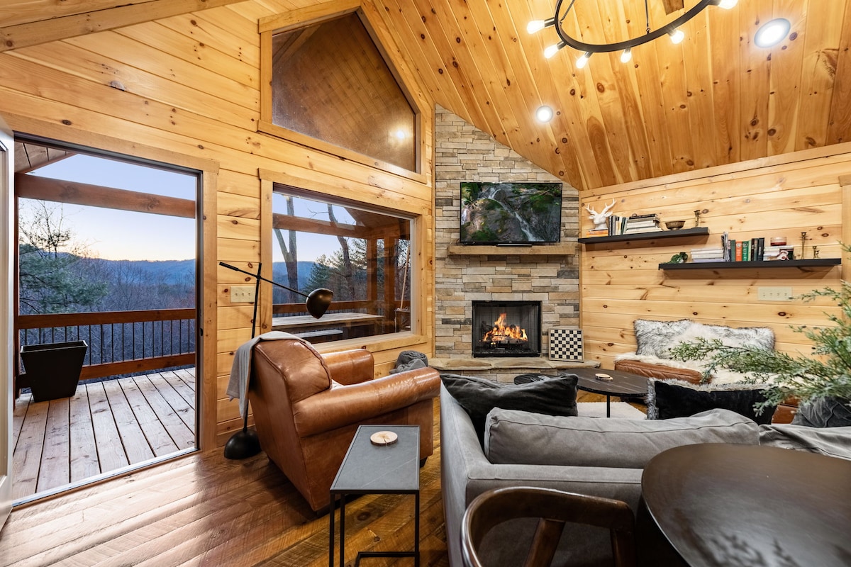 A cozy living space is highlighted by a stone fireplace and warm wooden walls. Large windows offer scenic views of the mountains. Comfortable seating options are arranged around a coffee table, while a wall-mounted television is visible. A deck is accessible through glass doors.