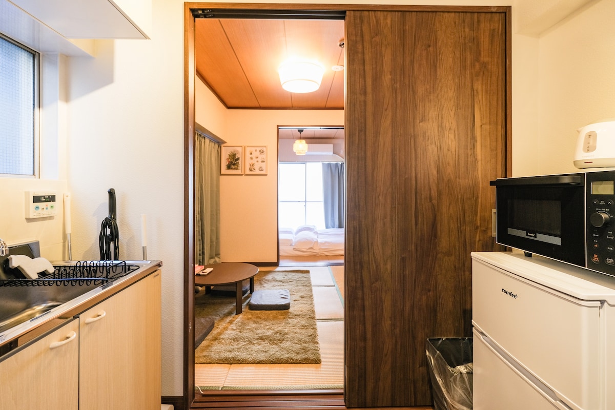 A view from the kitchen area into the living space is presented, featuring a sliding wooden door. The kitchen includes a sink and appliances, while the living area showcases a low table on a tatami mat, with bedding visible through the door.