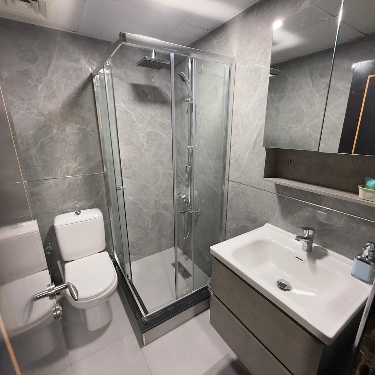 The bathroom features modern fixtures, including a glass-enclosed shower and a sleek vanity with a sink. A large mirror adds brightness, and gray tiles create a clean aesthetic. A toilet is positioned beside the shower, and complimentary toiletries are arranged neatly on the counter.