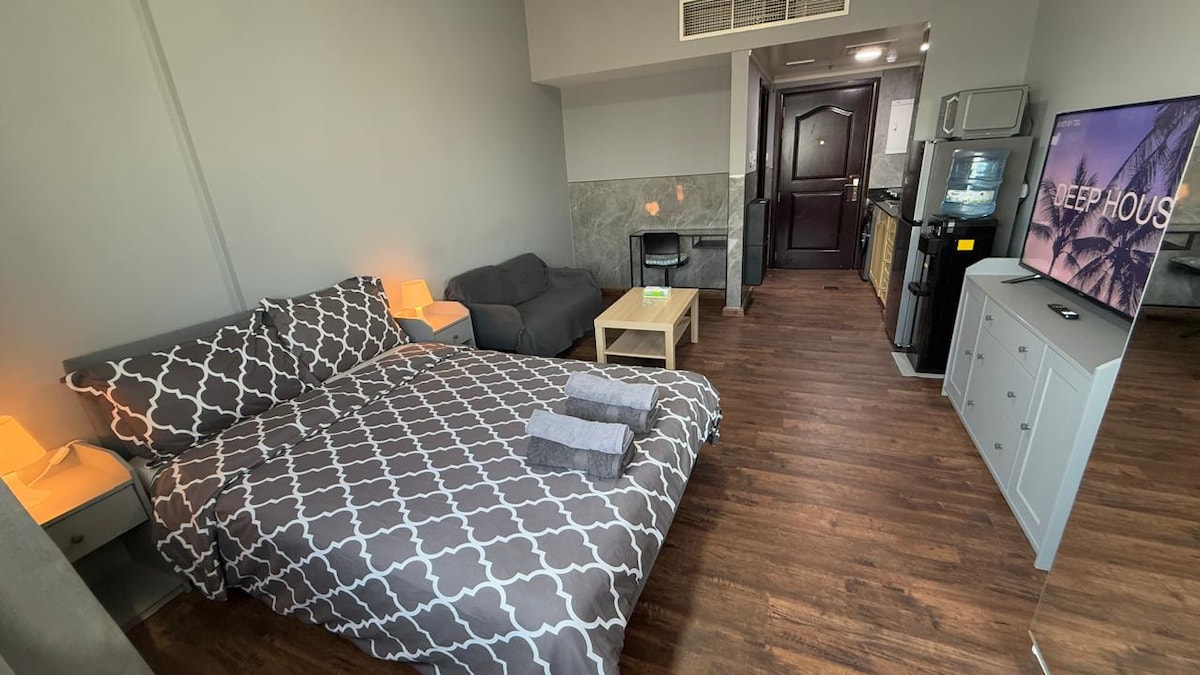 A well-arranged studio apartment features a spacious bed dressed in a patterned duvet, accompanied by two towels placed neatly on top. A small coffee table is positioned in front of a compact seating area, while a television stands on a console in the corner.