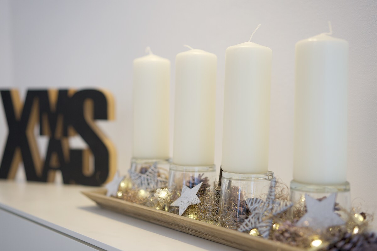 Four tall white candles are arranged on a decorative tray, accompanied by shimmering elements and small stars. A wooden sign with the text 'XMAS' adds a seasonal touch to the festive display.