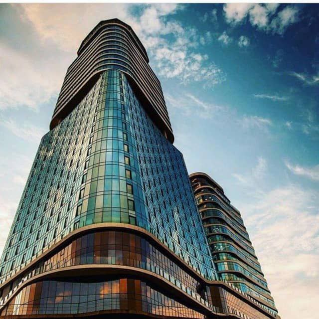 The modern architecture of the King David tower is highlighted from a low angle, showcasing its sleek glass facade and curvilinear design. The sky above features soft cloud formations, complementing the building's reflective surface.