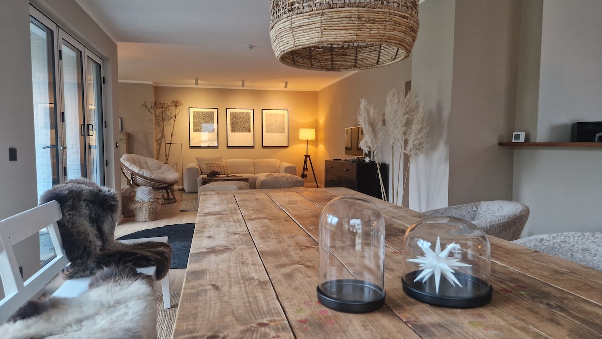 A dining area features a rustic wooden table at the forefront, accompanied by decorative glass cloches. In the background, a spacious living area is visible, highlighting a comfortable seating arrangement, framed artwork on the walls, and soft lighting that creates a calm ambiance.