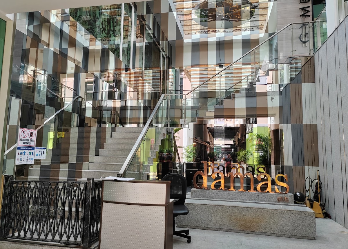 An inviting lobby area is showcased, featuring a welcoming reception desk. Glass walls reflect a modern design, creating a sense of openness. Stairs lead to upper levels adorned with greenery, while the 'Damas' sign is prominently displayed, enhancing the contemporary aesthetic of the space.