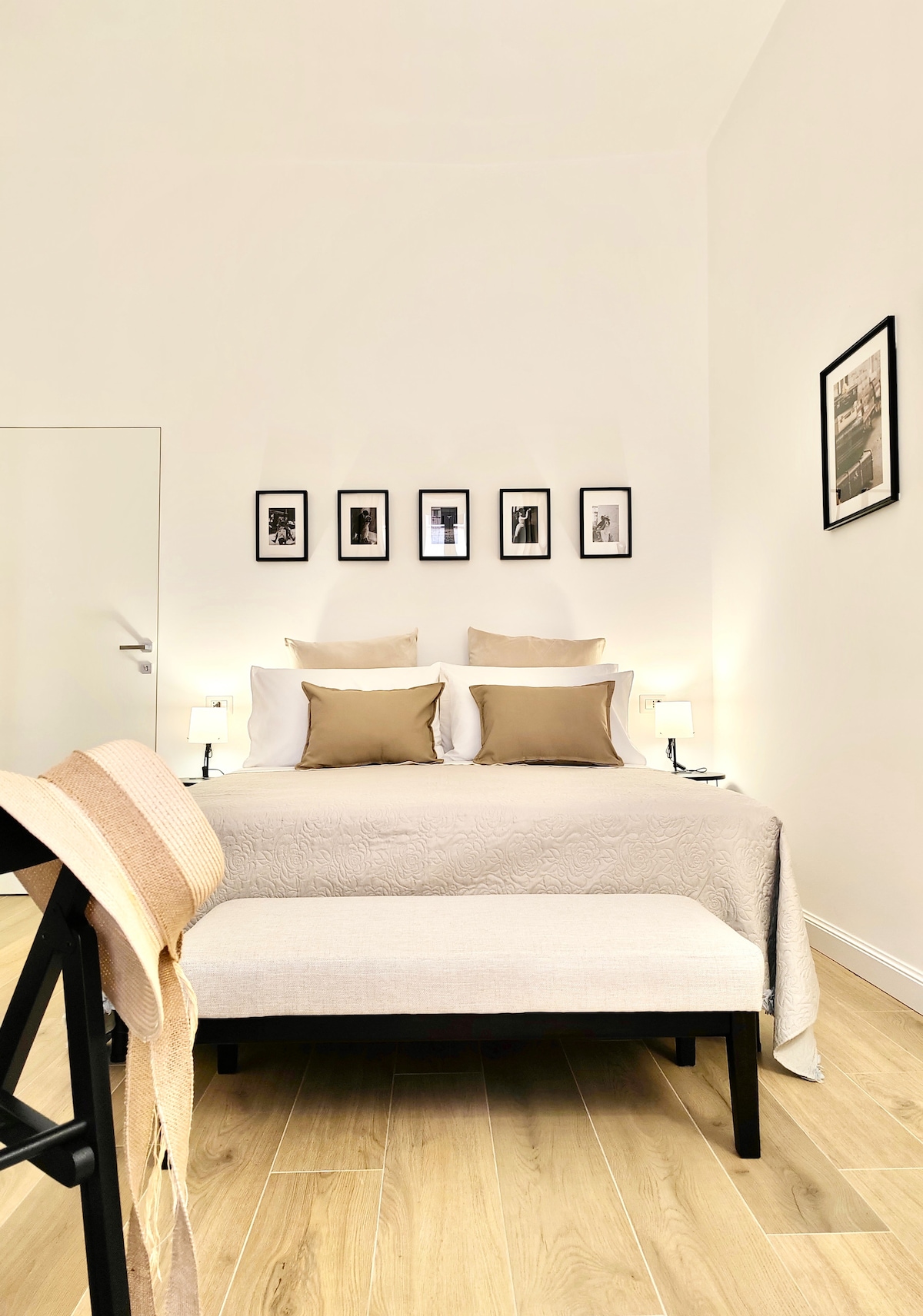 A serene bedroom is presented with a spacious bed adorned with decorative pillows and a soft quilt. Framed black-and-white photographs hang on the wall above. A stylish bench sits at the foot of the bed, while bedside lamps provide gentle lighting.