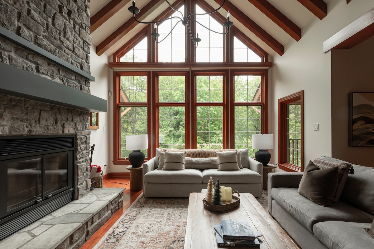 A spacious living area is highlighted by a stone fireplace and large windows that provide natural light and views of the surrounding greenery. Comfortable seating is arranged around a wooden coffee table, with decorative items placed on top.