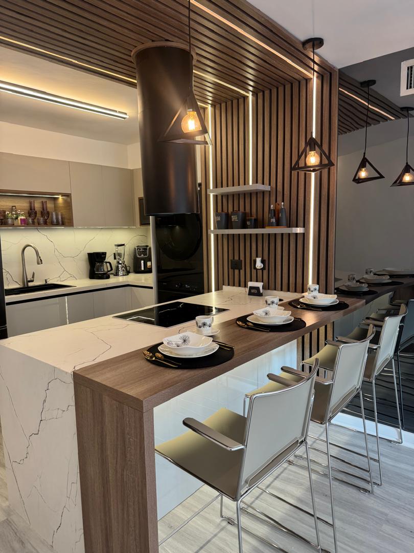 A modern kitchen is displayed, featuring a minimalist design with wooden accents and sleek cabinetry. The kitchen island is set with four place settings, and pendant lights illuminate the space, highlighting the elegant finishes and ample counter space. Appliances are seamlessly integrated into the design.