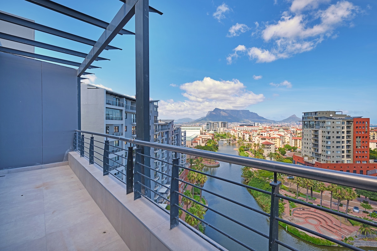 A spacious private balcony offers sweeping views of Table Mountain and the surrounding cityscape. The railing is composed of metal slats, reflecting a modern design. The scene includes urban buildings, greenspaces, and a tranquil waterway that enhances the natural beauty of the area.