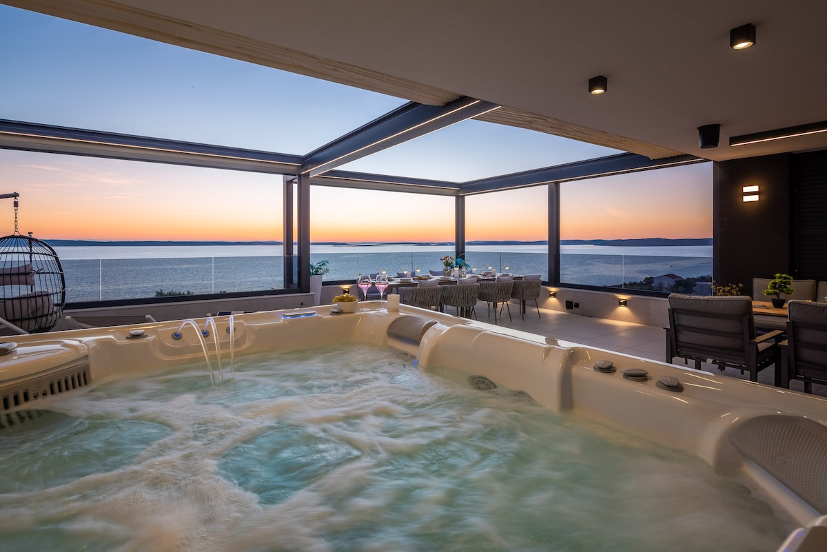 A private spa area features a spacious hot tub, surrounded by glass walls that provide an unobstructed view of the sea. Outdoor seating is visible in the background, with a dining setup set against a colorful sunset, enhancing the tranquil ambiance.
