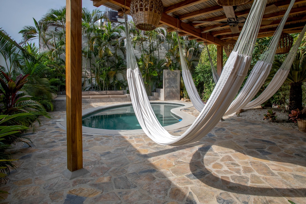 A serene outdoor area features a stone patio adorned with hammocks hanging from a wooden structure. A clear pool is positioned nearby, surrounded by lush greenery and tropical plants. Natural light enhances the peaceful ambiance in this inviting space.