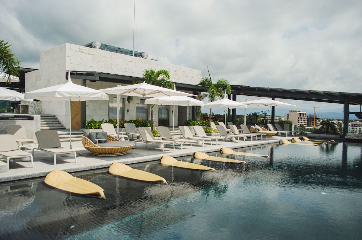 An expansive infinity pool is surrounded by sun loungers, each paired with white umbrellas. Lush greenery is positioned along the pool's edge, while a stylish stone structure sits above, featuring a modern design and outdoor spaces for relaxation.