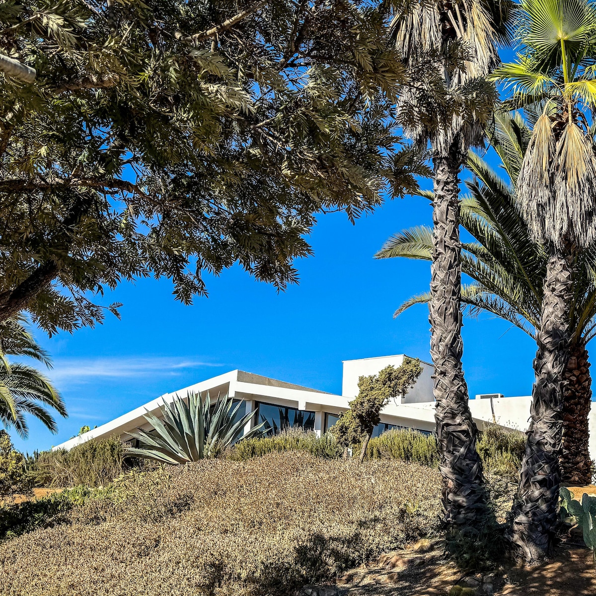 A modern villa is situated amidst lush greenery and palm trees, showcasing a striking architectural design with sloped roofing. Clear blue skies provide a bright backdrop, enhancing the serene outdoor atmosphere surrounding the property.