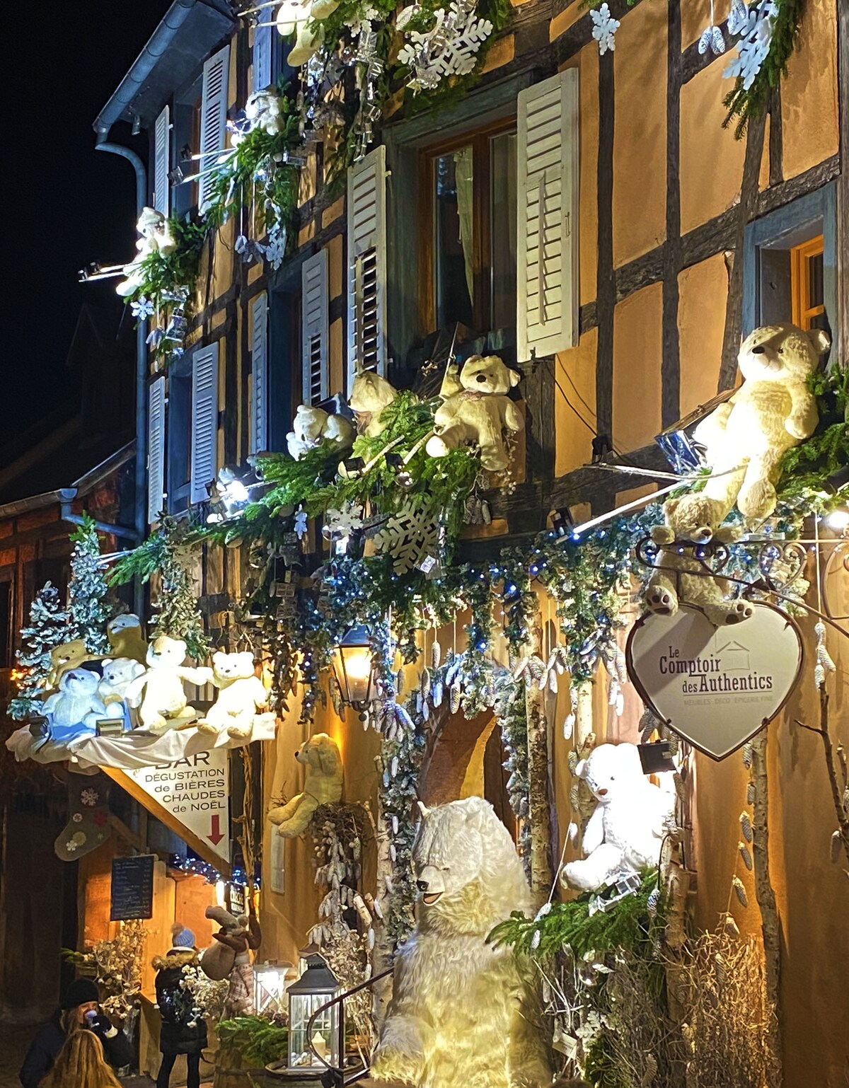 The facade of a charming building is adorned with festive decorations, featuring white teddy bears and greenery. Soft lights illuminate the scene, highlighting the wooden beams and shutters. A heart-shaped sign is visible amidst the decor, creating a welcoming and festive atmosphere.