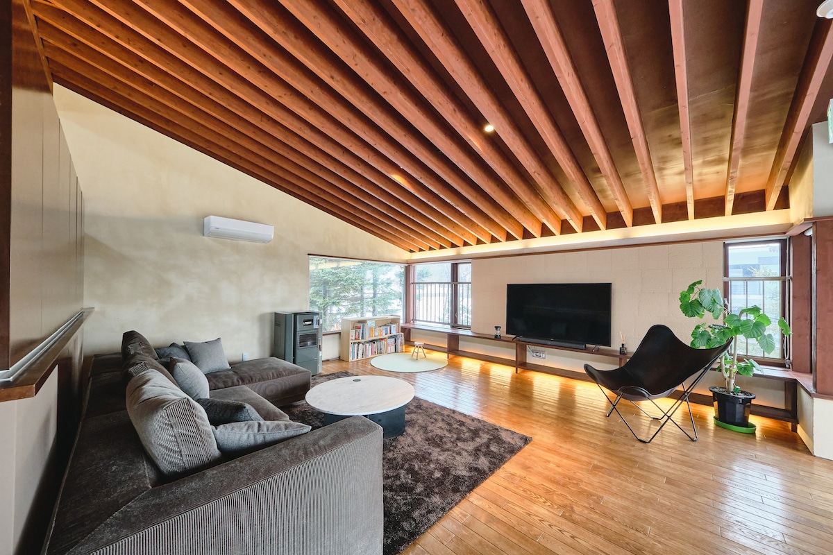 The living area features a large, comfortable sectional sofa facing an entertainment system mounted on the wall. A round coffee table is placed in front of the sofa, with a textured rug covering the wooden floor. Large windows allow natural light to illuminate the space.