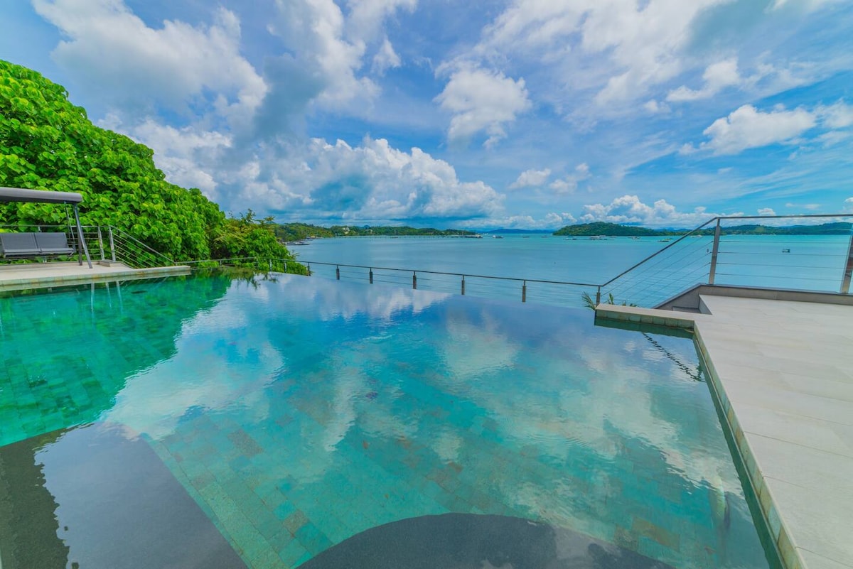 An infinity pool seamlessly blends with the ocean, reflecting the sky's vibrant blue and fluffy white clouds. Lush greenery surrounds the area, enhancing the natural ambiance. The tranquil water invites relaxation, while a sleek railing provides a clear view of the serene seascape.