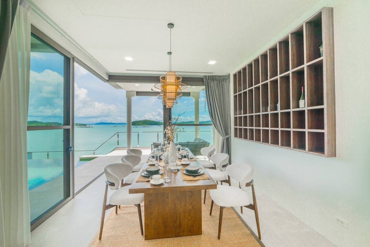 A modern dining area features a wooden table surrounded by six chairs, set against large glass doors offering views of the pool and ocean. Soft natural light fills the space, complemented by decorative shelving on the wall displaying select items.