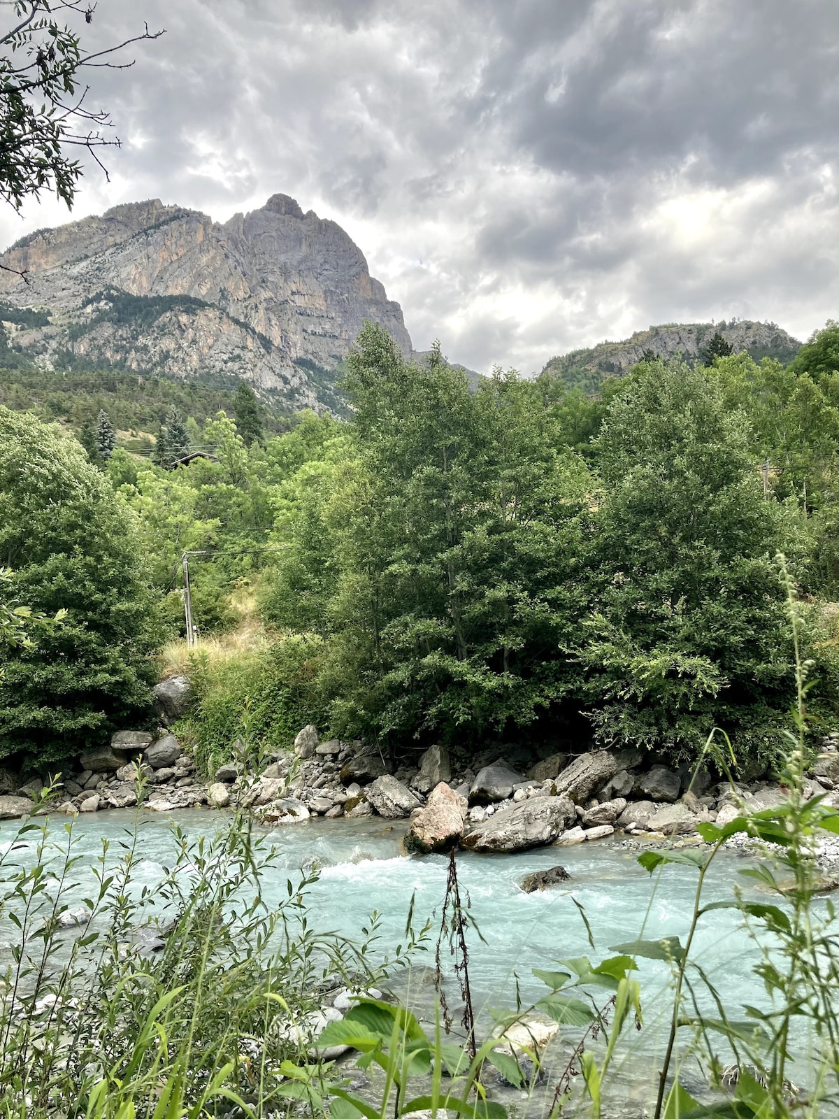 Lush greenery surrounds a gentle river, with smooth stones visible along its edge. Towering mountains rise in the background under a cloudy sky, creating a serene natural landscape. The water's light blue hue adds to the tranquil atmosphere.