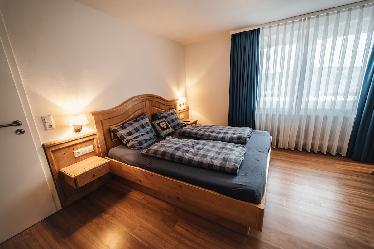 A well-lit bedroom features a wooden bed with two separate mattresses, layered with patterned bedding. Nightstands with lamps are positioned on either side. Natural light filters through sheer curtains, complementing the warm wood floors and creating a cozy ambiance.