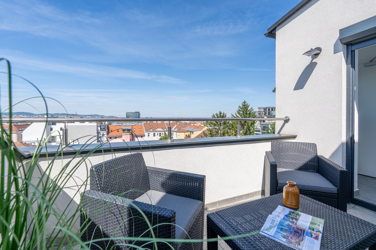 A private balcony displays modern outdoor furniture, including two chairs and a small table. Lush greenery partially frames the space, complemented by a scenic view of the surrounding rooftops and clear blue skies, creating a serene environment for relaxation.