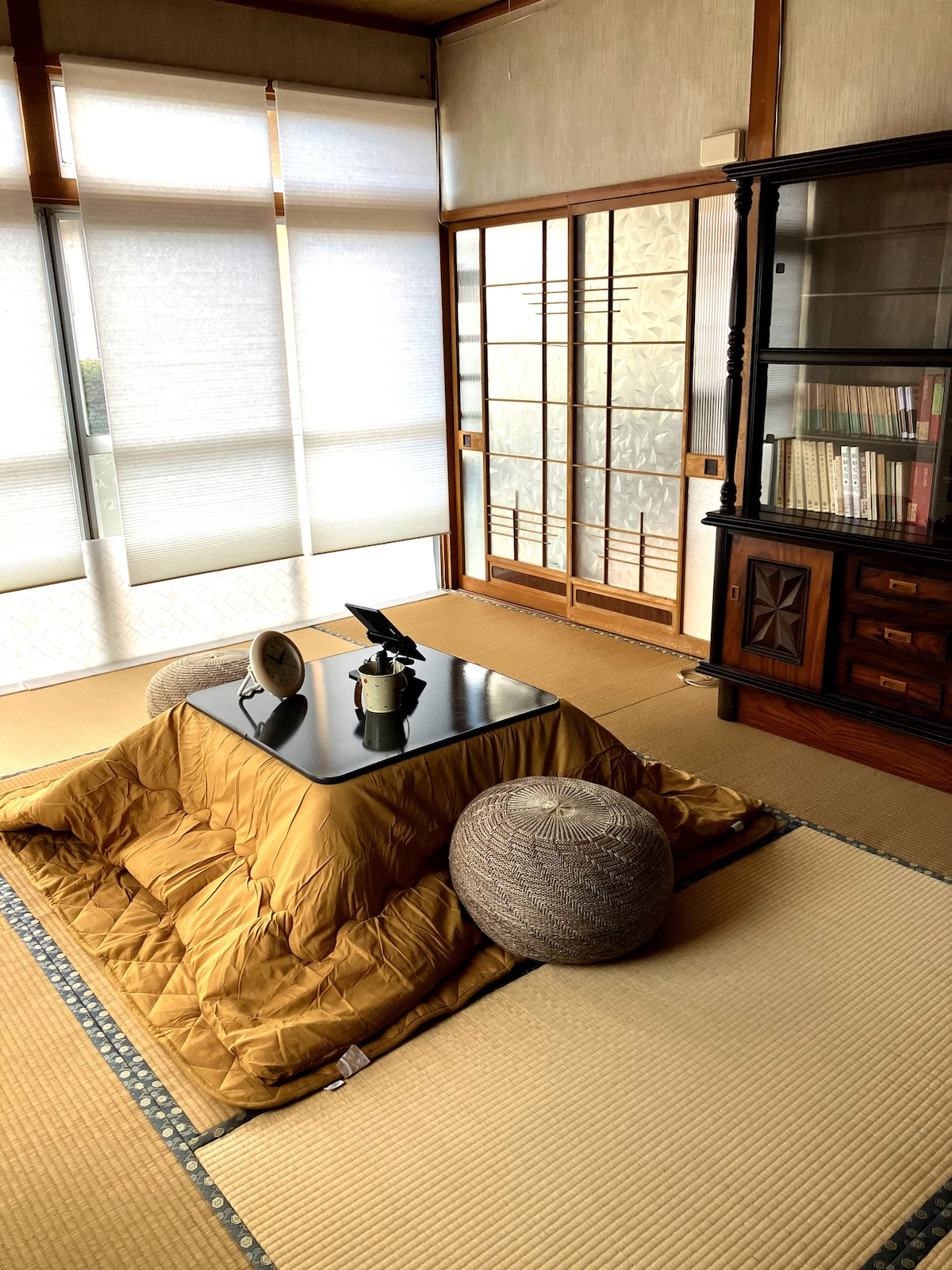 A tatami-floored room features a low table surrounded by poufs. Natural light filters through partially closed shoji screens, illuminating the warm tones of the space. A wooden cabinet displays books and decorative items in the background.