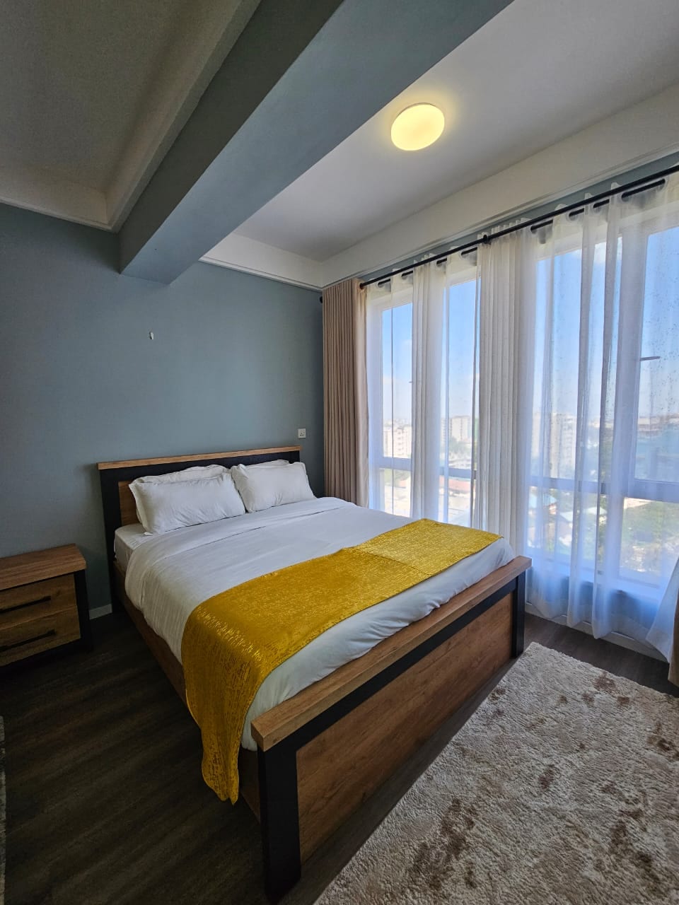A spacious bedroom is illuminated by natural light streaming through large curtains. A well-made queen-sized bed is dressed in crisp white linens, with a soft yellow throw blanket draped across the foot. Nightstands complement the warm wood tones of the bed.