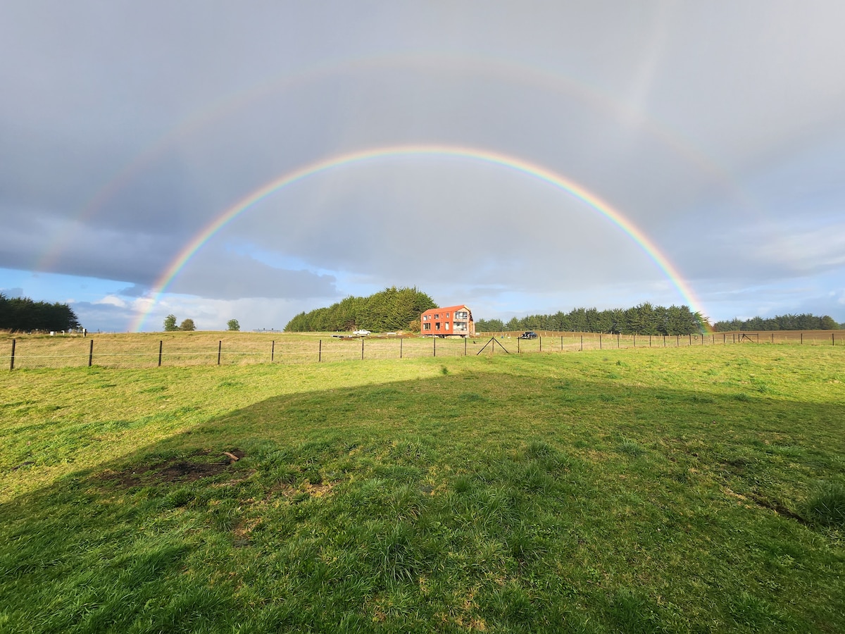 A rainbow arches across the sky above a green, open field. A simple, red house is visible in the distance, surrounded by trees and a fence line. The landscape offers a serene and natural setting.