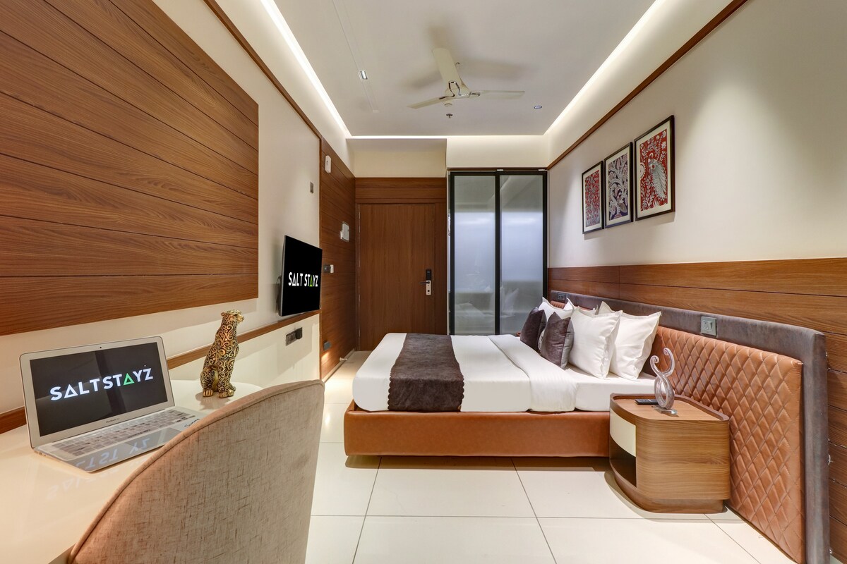 A modern room is depicted, featuring a spacious bed with neutral-toned bedding. A desk with a laptop is positioned next to a decorative item. Two framed artworks adorn the wall above the headboard, and large sliding doors provide access to the balcony.