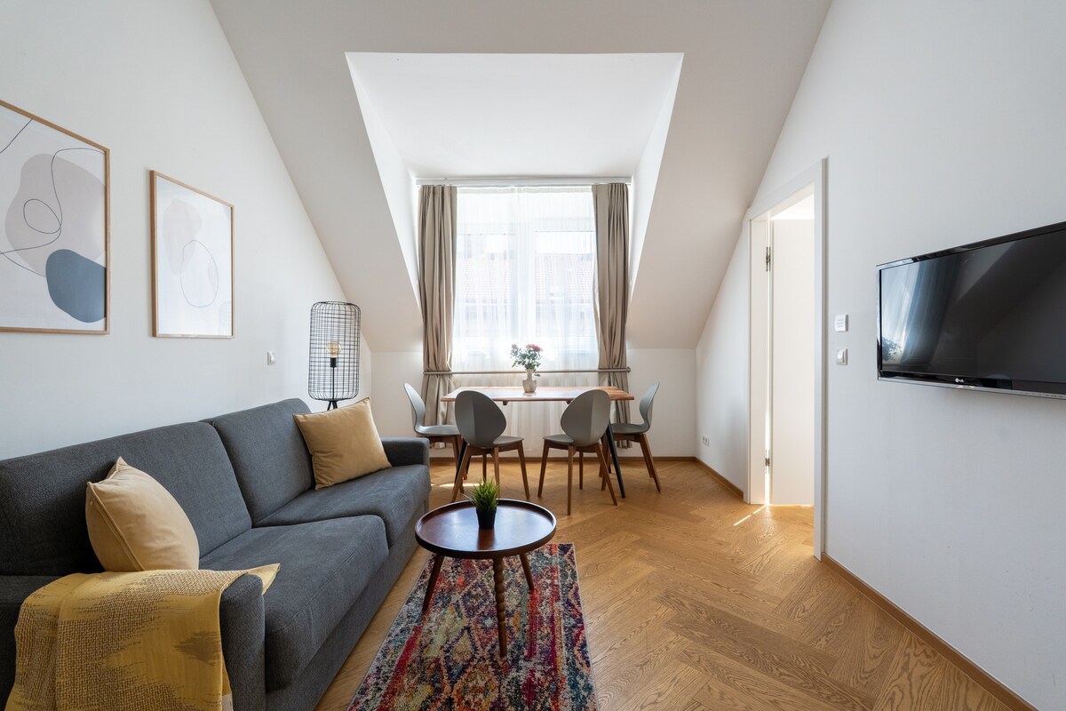 A modern living area features a gray sofa and a round coffee table placed on a colorful patterned rug. Natural light enters through a large window, while a dining table with four chairs is positioned nearby. Wall art adds subtle decoration to the space.