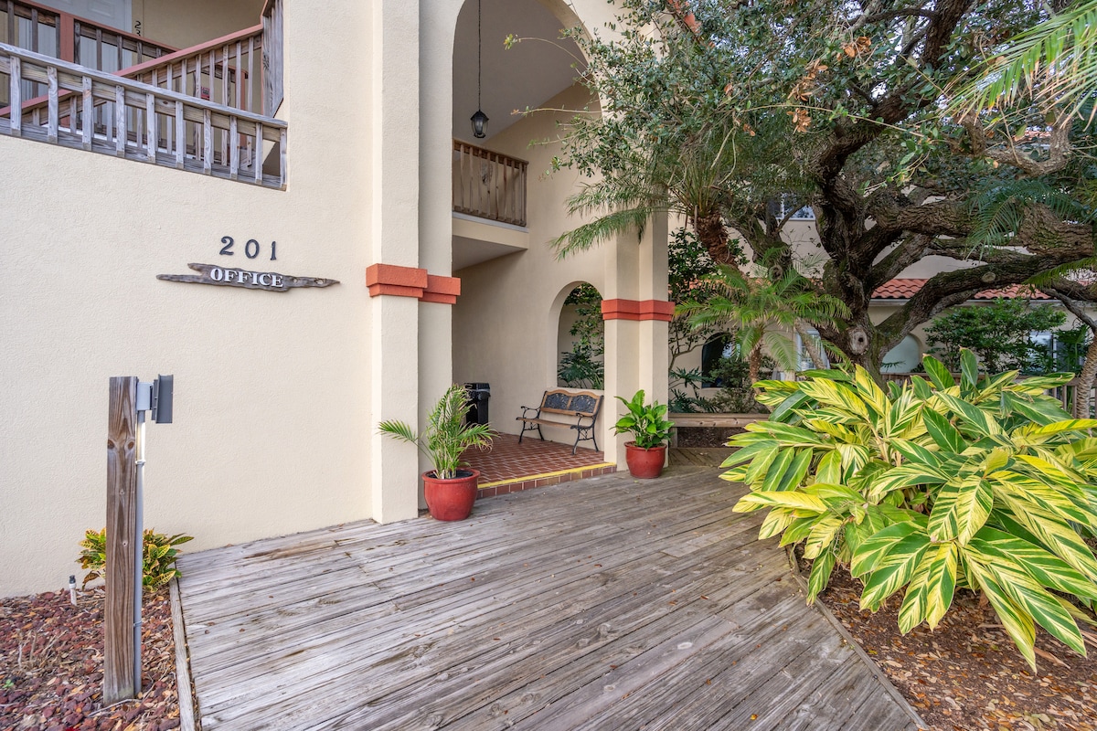 The entrance to the office is framed by lush greenery and potted plants, with wooden decking leading up to the arched entryway. A wooden bench sits beside the entrance, providing a place to relax in the shaded area under a large tree.
