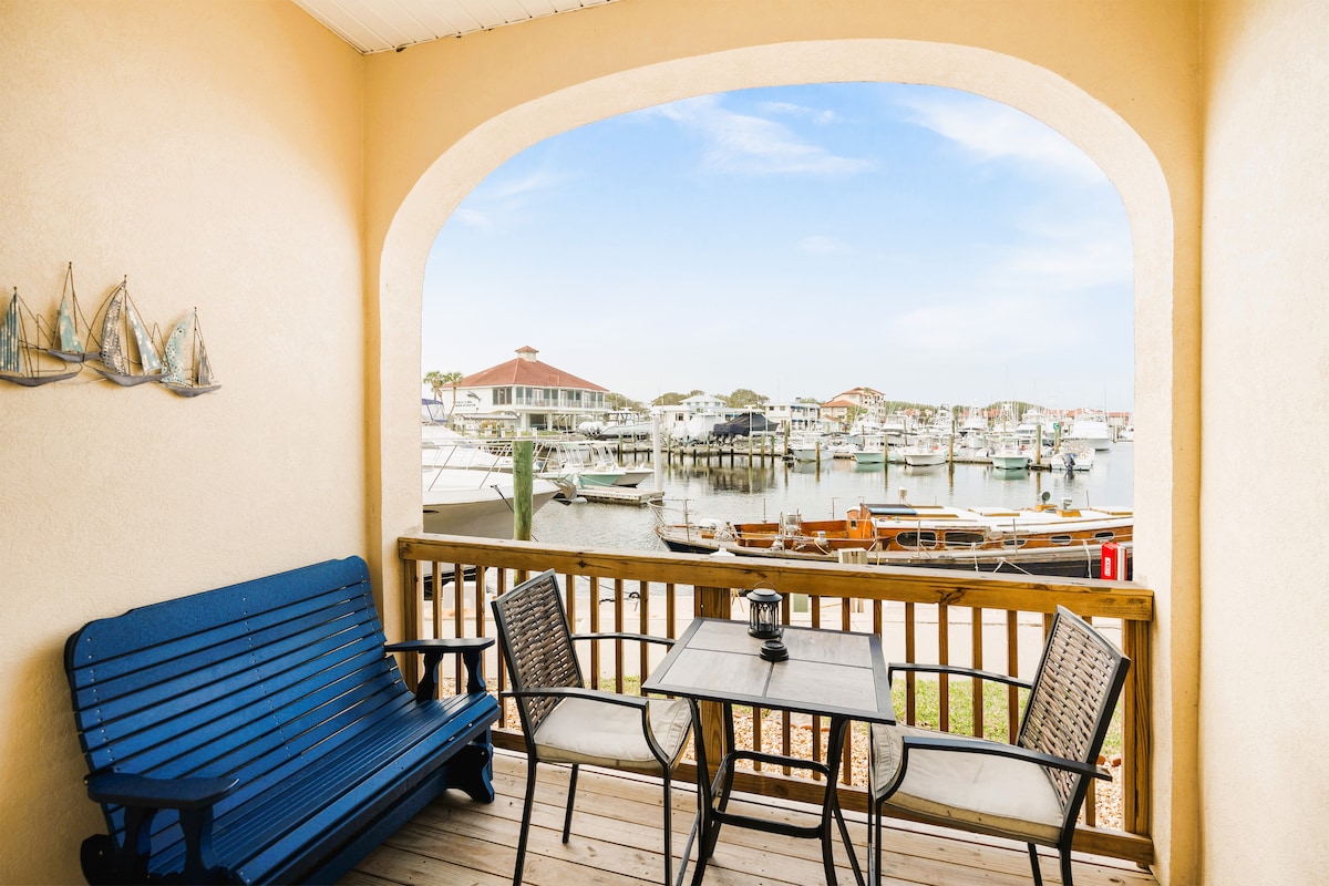 A charming patio space features a small table with two chairs and a blue swing. The view of the marina is framed by arches, showcasing boats and waterfront activity, providing a serene outdoor setting for relaxation.