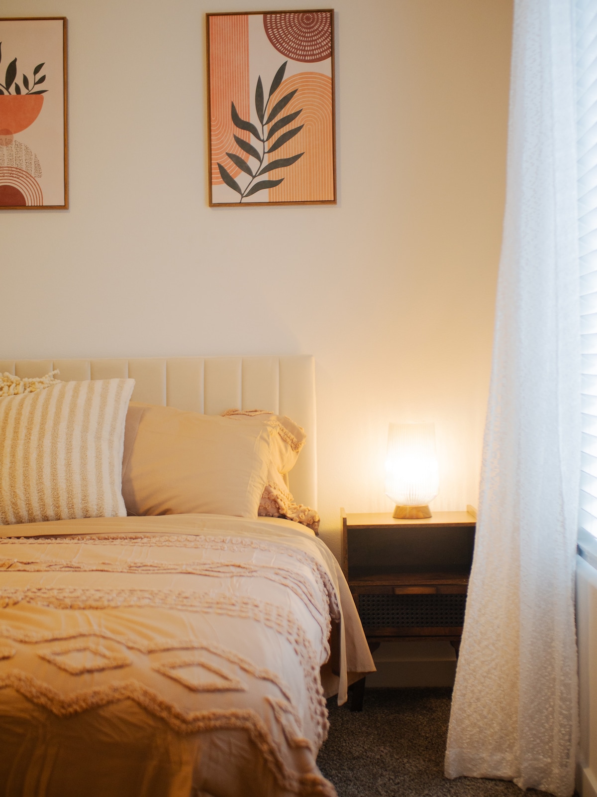 A cozy bedroom features a spacious bed adorned with soft beige linens and decorative pillows. A minimalist nightstand holds a warm lamp, casting a soft glow. Two framed artworks adorn the wall, and a sheer curtain gently filters the natural light from the window.