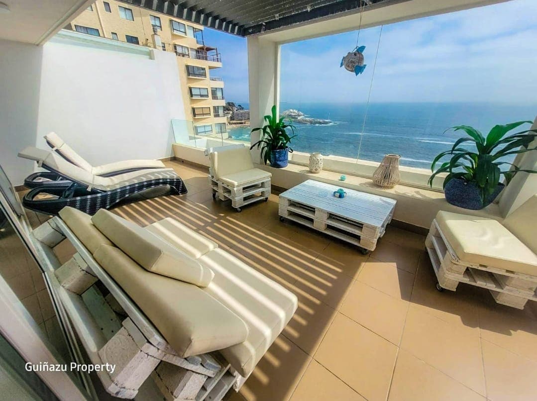 A spacious terrace offers comfortable seating arrangements with loungers and a coffee table. Panoramic views of the ocean can be observed through large glass windows, while potted plants add a touch of greenery to the space.