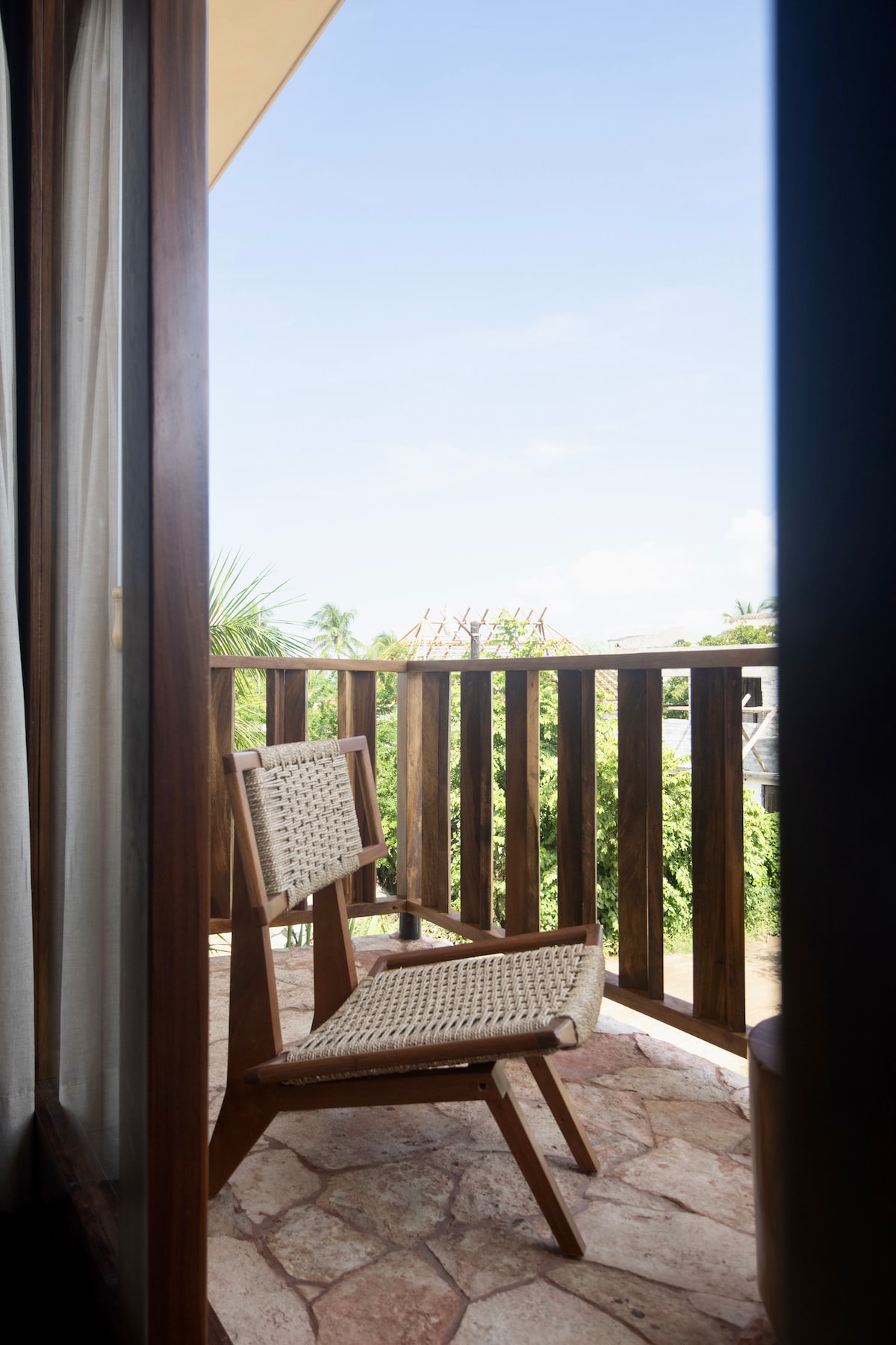 A wooden balcony features a woven armchair, overlooking lush greenery and a clear blue sky. The natural stone floor adds a rustic touch, while the wooden railing offers an open view, allowing for a serene outdoor space.