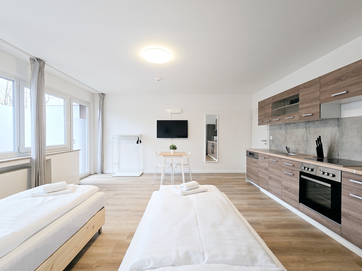 A studio apartment features two separate beds with simple white linens. A modern kitchen with wooden cabinetry is visible, alongside a dining table set for two. Large windows allow natural light to fill the space, and a television is mounted on the wall.