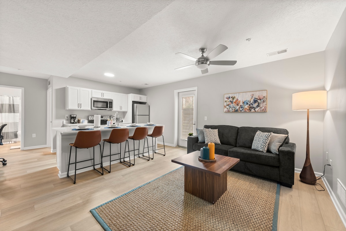 An open concept living area is showcased with hardwood flooring, featuring a comfortable sofa and a wooden coffee table. A well-equipped kitchen is visible with modern appliances and bar seating. A painting adorns the wall, and natural light enters through the windows, enhancing the space.