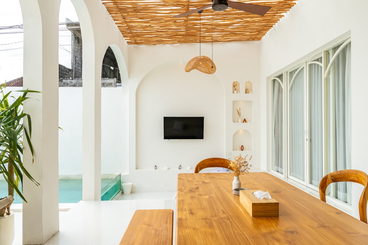 A spacious dining area features a large wooden table surrounded by curved chairs. A fan hangs from the ceiling above, and a television is mounted on the wall. Natural light filters in through arches, with a glimpse of the private pool visible in the background.