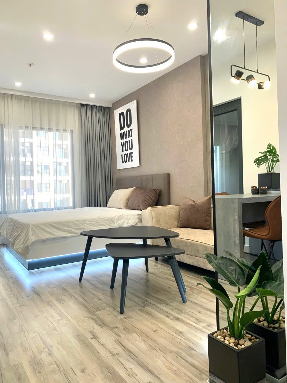 A modern studio apartment is presented, featuring a spacious bed with neutral bedding and accented by two brown pillows. A comfortable sofa and a small black table are included. Natural light enters through the large window, framed by sheer drapes, enhancing the interior's contemporary feel.