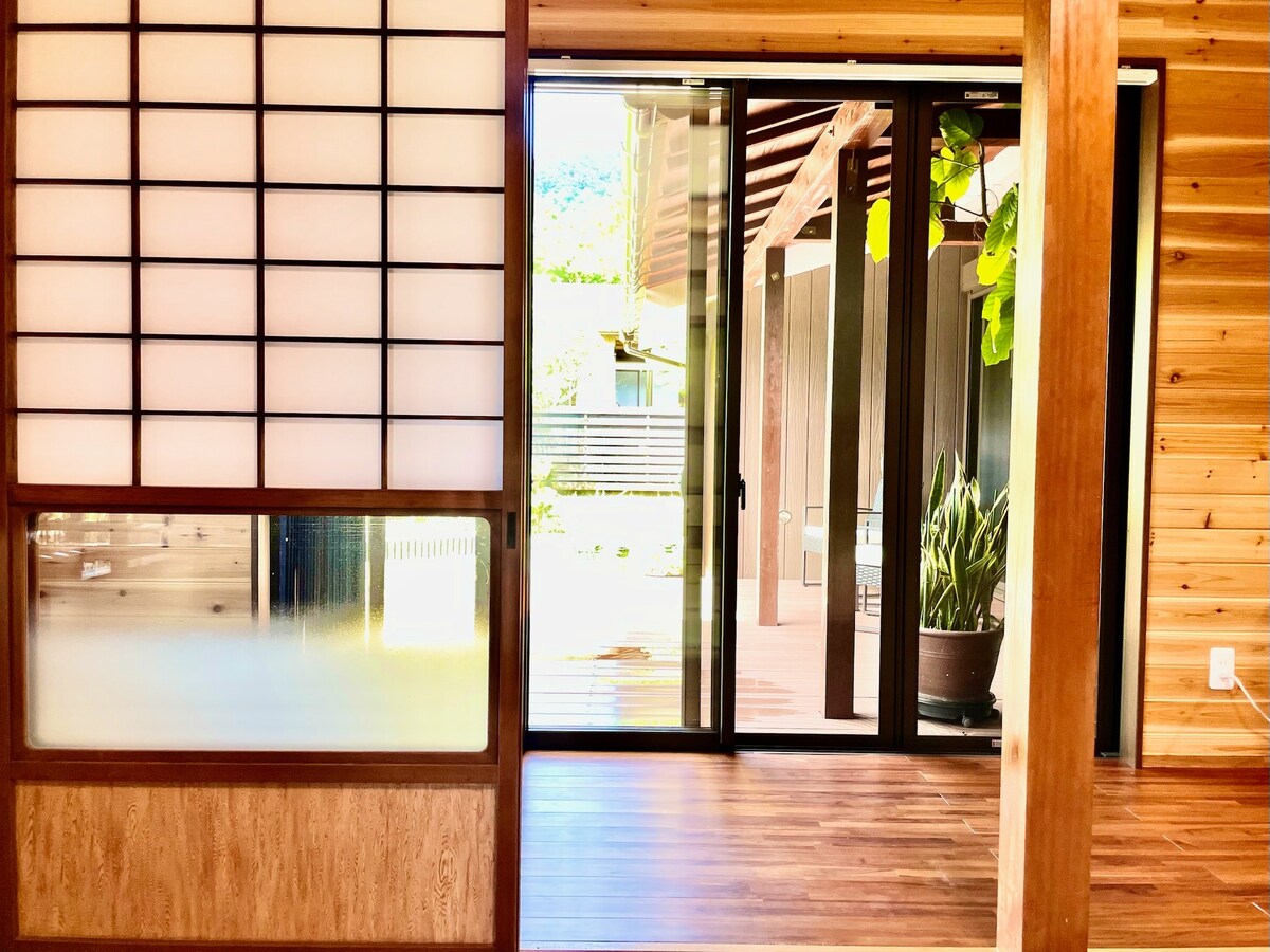 An entrance area is featured, connecting the interior to the outdoor space. Sliding glass doors provide a view of the garden, while wooden paneling and shoji screens create a warm and inviting aesthetic. Natural light filters through, enhancing the tranquil atmosphere.