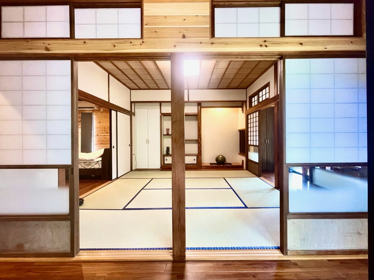 A traditional Japanese-style room divided by shoji screens features tatami mat flooring and minimalist decor. Natural wood accents and sliding doors create a light, airy atmosphere, while ample wall space allows for decorative touches. A serene ambiance is established by the symmetrical layout.